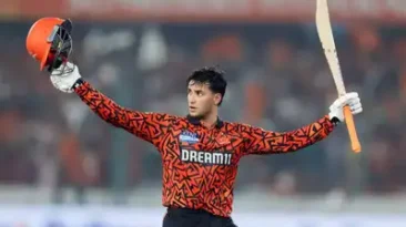 Abhishek Sharma of Sunrisers Hyderabad celebrates scoring 141 runs in IPL 2025, holding his bat high in a floodlit stadium with fireworks in the sky and a cheering crowd in the background.