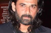 Portrait of Indian actor Mukul Dev smiling, wearing a formal suit, in a dimly lit background.